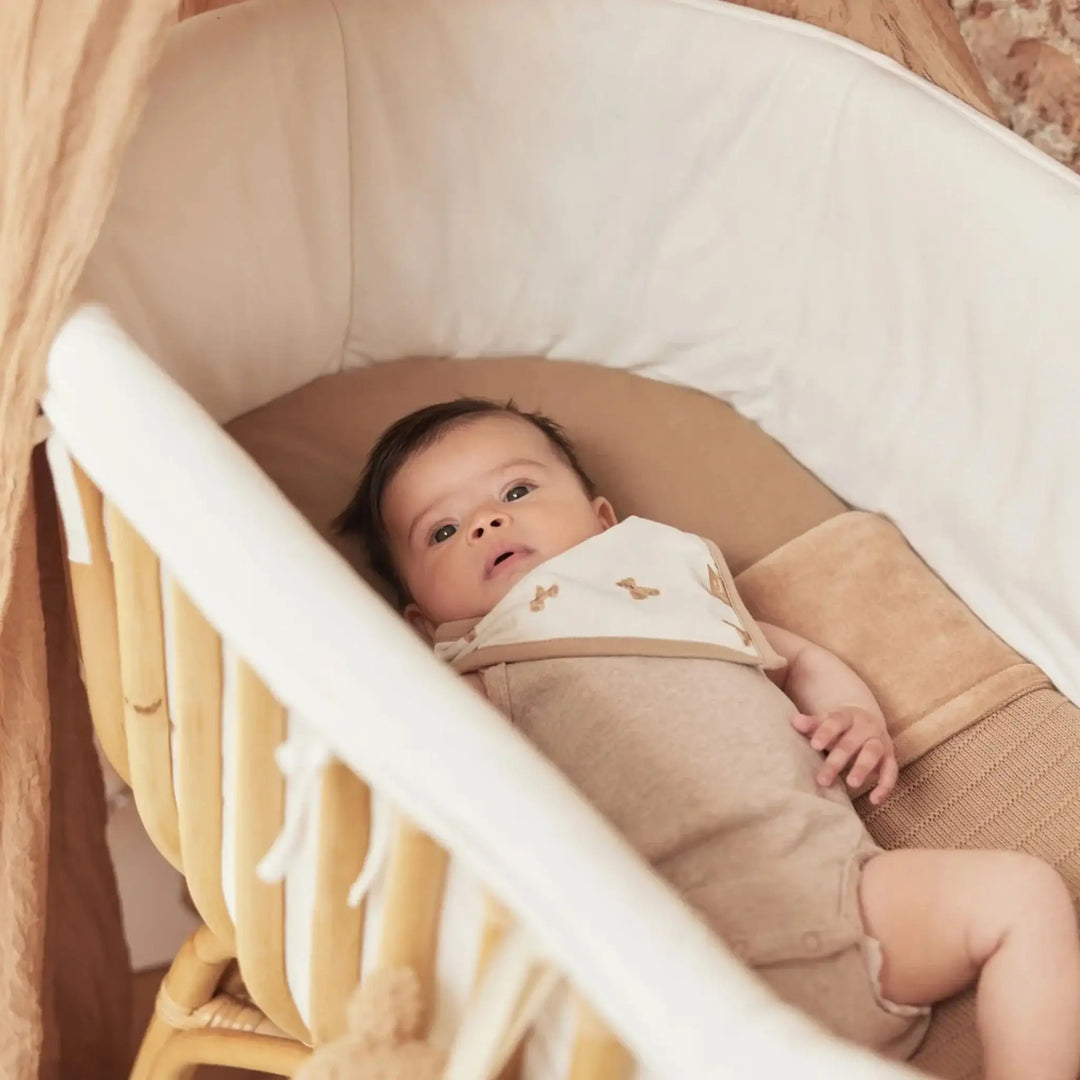 Baby in beige romper liggend in rotan wieg met zachte dekens, neutrale kinderkamer