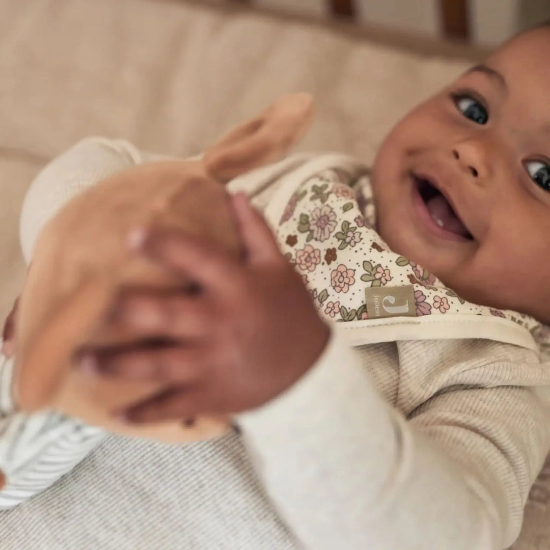 Lachende baby met bloemen slabbetje en zachte knuffel op neutraal beddengoed