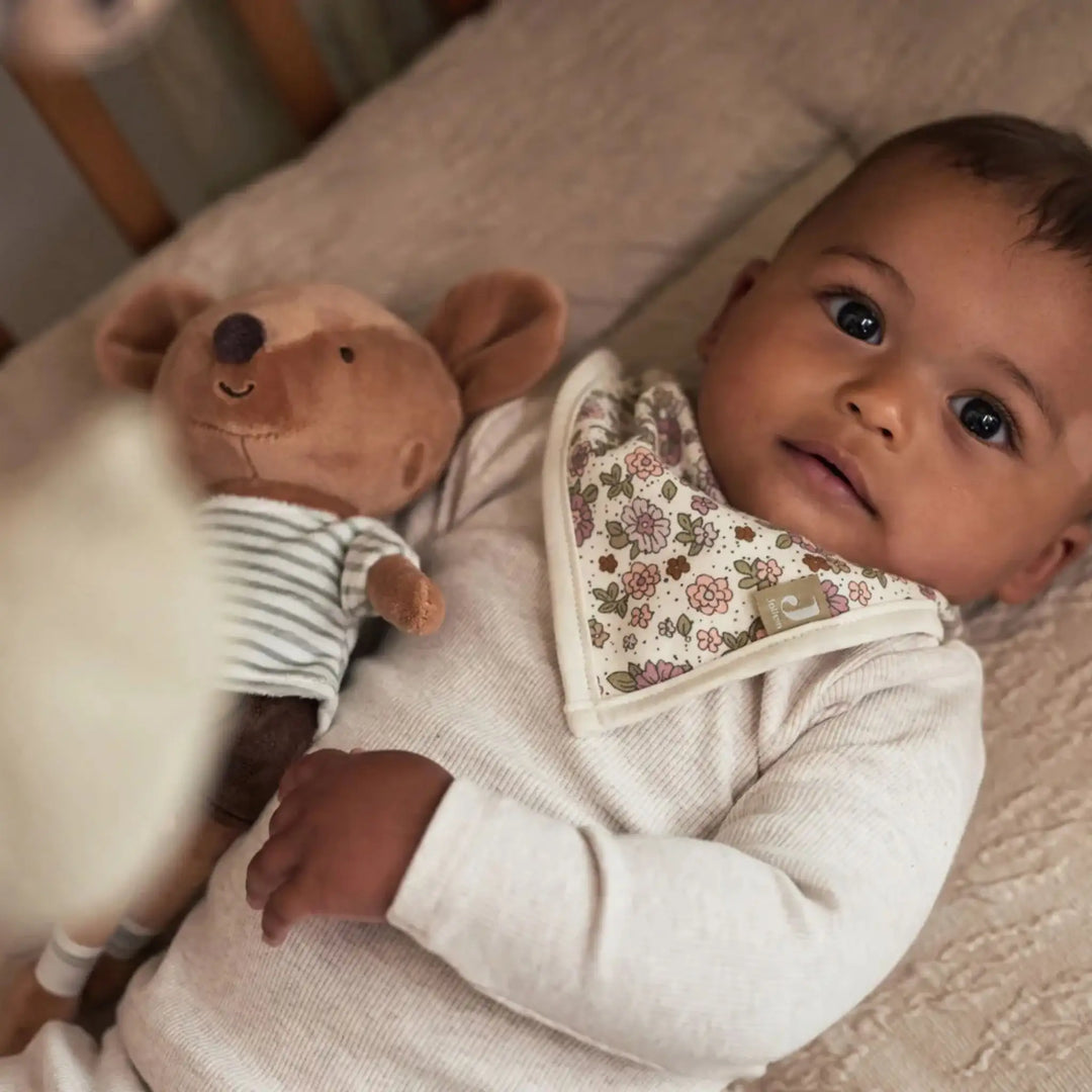 Baby in beige rompertje met bloemen slabbetje en knuffelmuis op bed, kindercollectie.